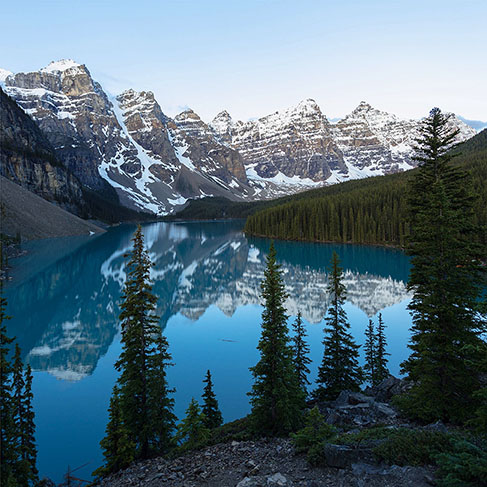 Pine trees near lake reflecting mountains under a clear sky