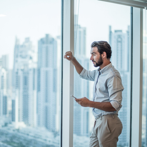 Businessman with smartphone staring through window with skyscraper view